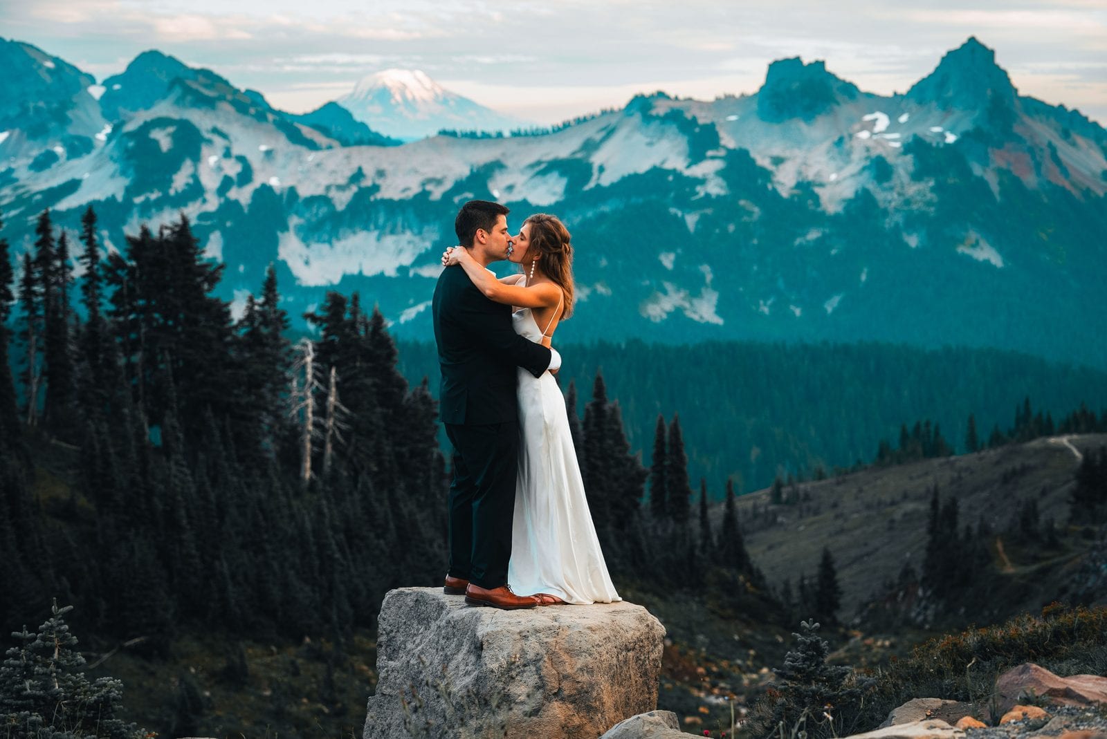 The couple embracing on a rock with the mountains behind
